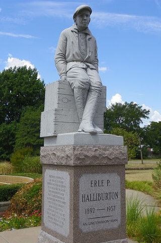 Erle P. Halliburton in work clothes and boots casually sitting on two wooden crates, a life-size statue on pink granite base.