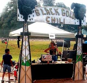 A woman at her "Black Gold Chili" stand, which has oil-gushing derrick props on each side. 